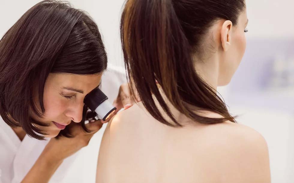 A female skin doctor closely inspects a mole on the left shoulder of a female patient using a dermatoscope